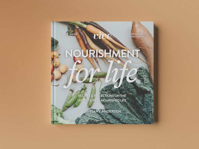 Nourishment for Life cookbook by Brittany Andersen, lying on a warm tan background with a cover photograph of fresh carrots, peas, kale, and onions.